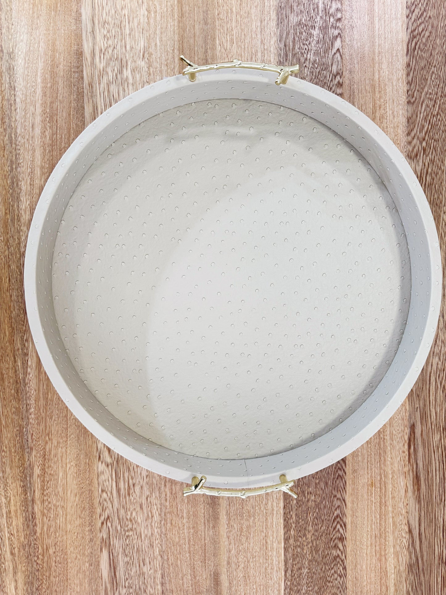 Vista superior de bandeja redonda en polipiel taupe con asas doradas de acero inoxidable, acabado premium